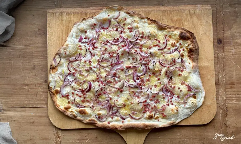 Frisch gebackener Flammkuchen mit knusprigem Sauerteigboden aus Weizen-Anstellgut, belegt mit Schmand, Schinkenwürfeln und Gruyère-Käse.